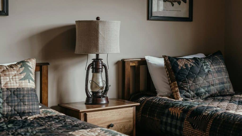 A rustic lamp sits on a nightstand between two beds with patterned quilts.