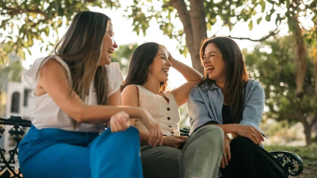 Three happy women sit on a bench, laughing and enjoying each other's company outdoors.
