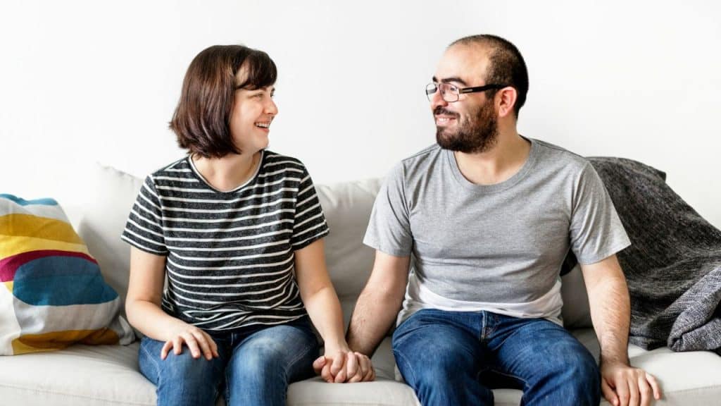 A happy couple sits on a couch, holding hands and smiling at each other.