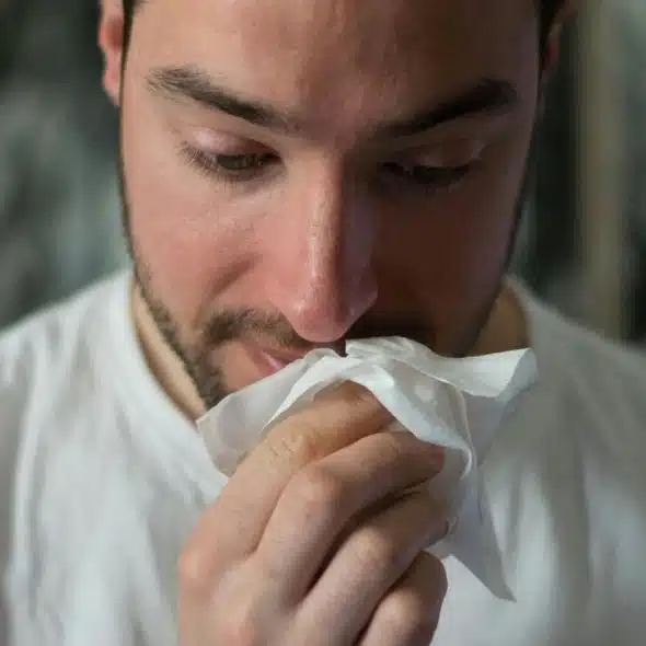 A close-up of a bearded man blowing his nose into a crumpled tissue.