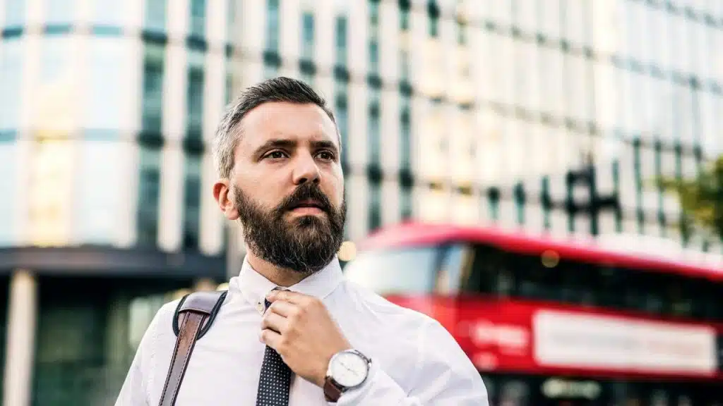 A bearded businessman adjusting his tie with a red bus and glass building behind him.