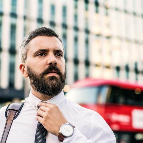 A bearded businessman adjusting his tie with a red bus and glass building behind him.