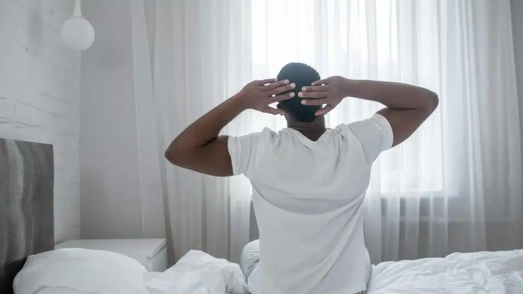 A man stretching on a bed facing a bright window.