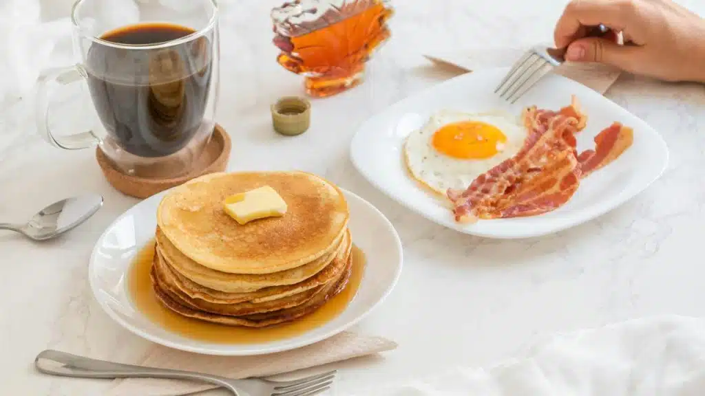 A stack of buttered pancakes with syrup next to bacon, a fried egg, and a cup of coffee.