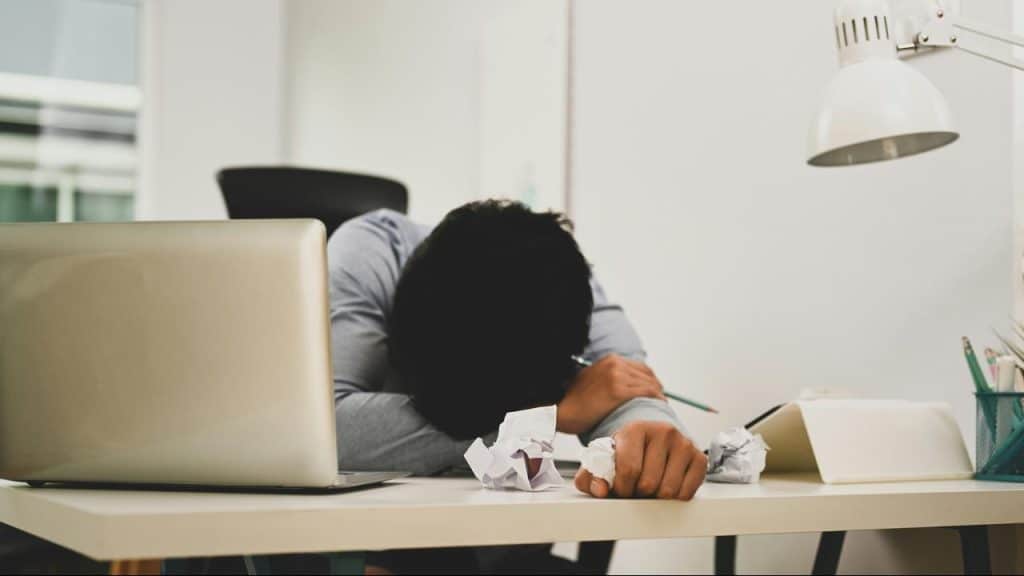 A man slumped over a desk scattered with crumpled papers.