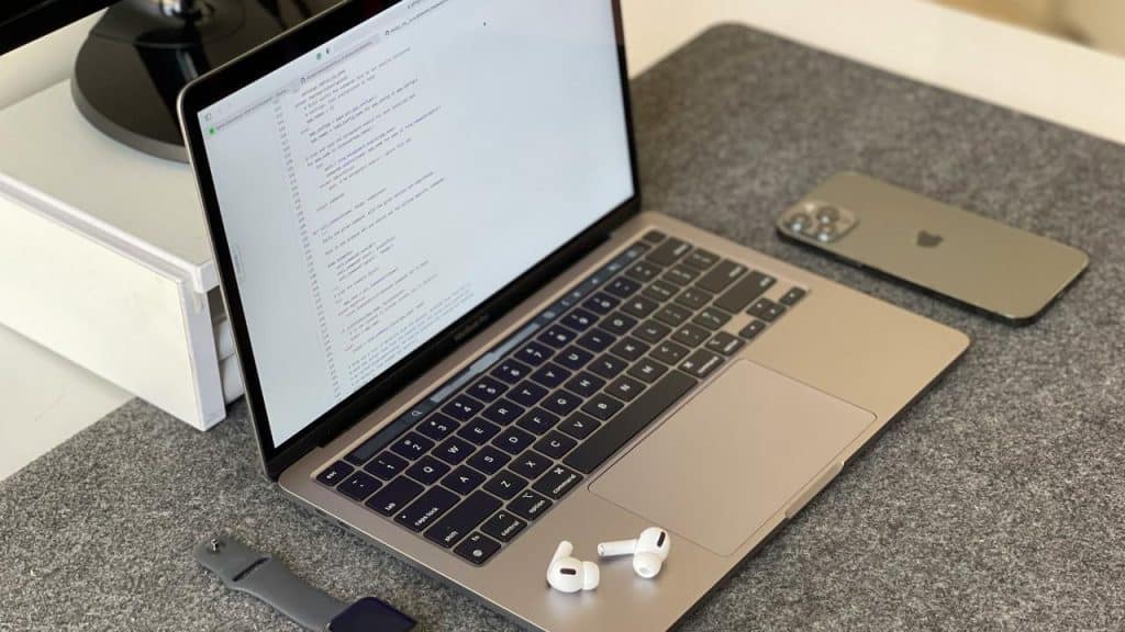 A laptop showing code, with earbuds, a smartwatch, and a smartphone beside it.
