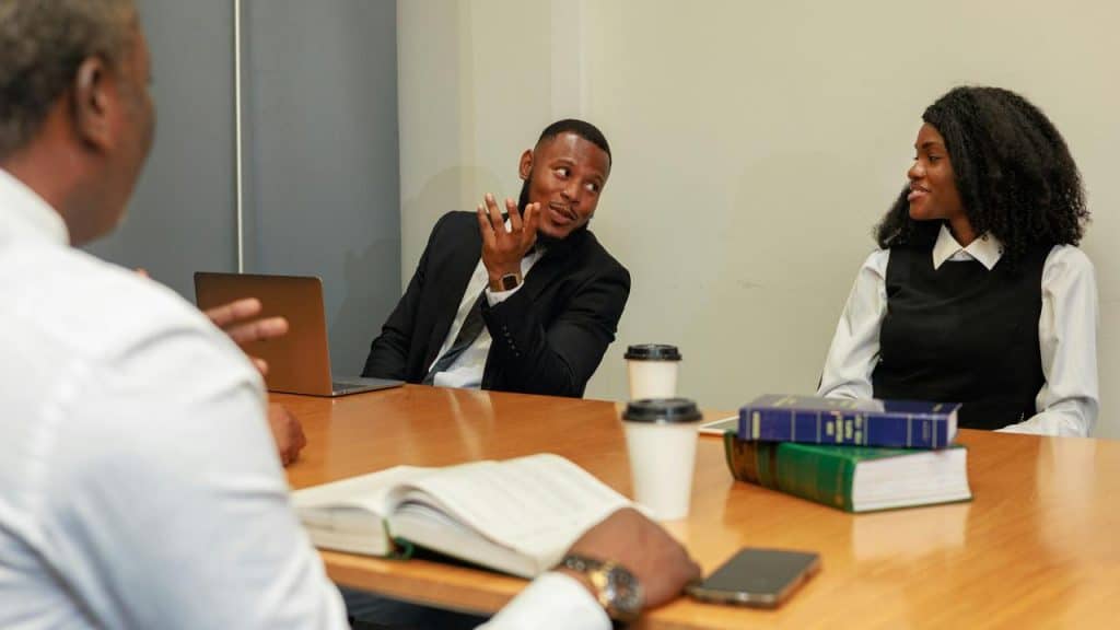 A group of three professionals in a meeting around a table with coffee cups, books, and a laptop.