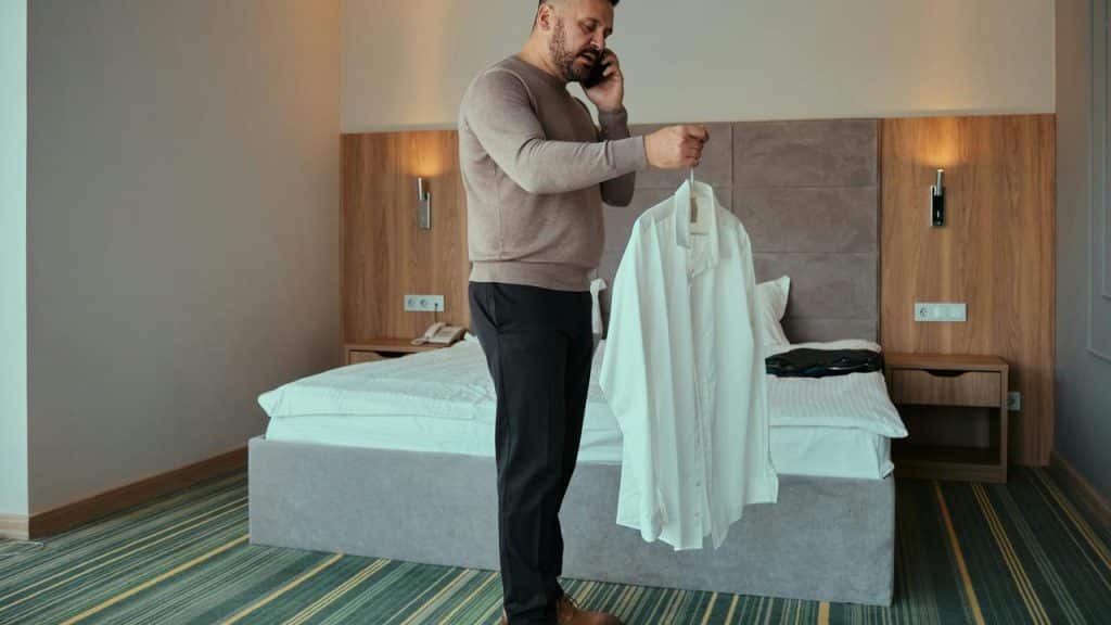 A man on the phone holding a white dress shirt on a hanger in a bedroom.
