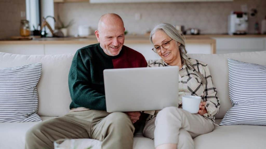 An older couple sitting on a living room couch, smiling as they look at a laptop together.