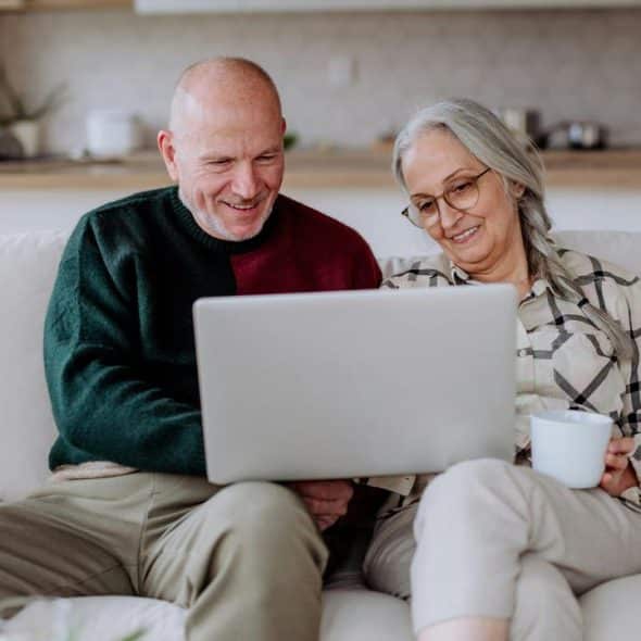 An older couple sitting on a living room couch, smiling as they look at a laptop together.