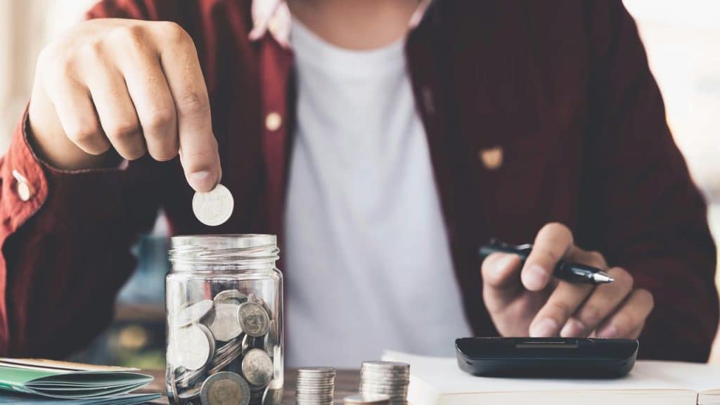 A person placing a coin into a jar of savings beside a calculator.
