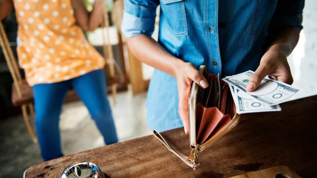 A person pulling dollar bills from a wallet over a wooden counter with a service bell nearby.