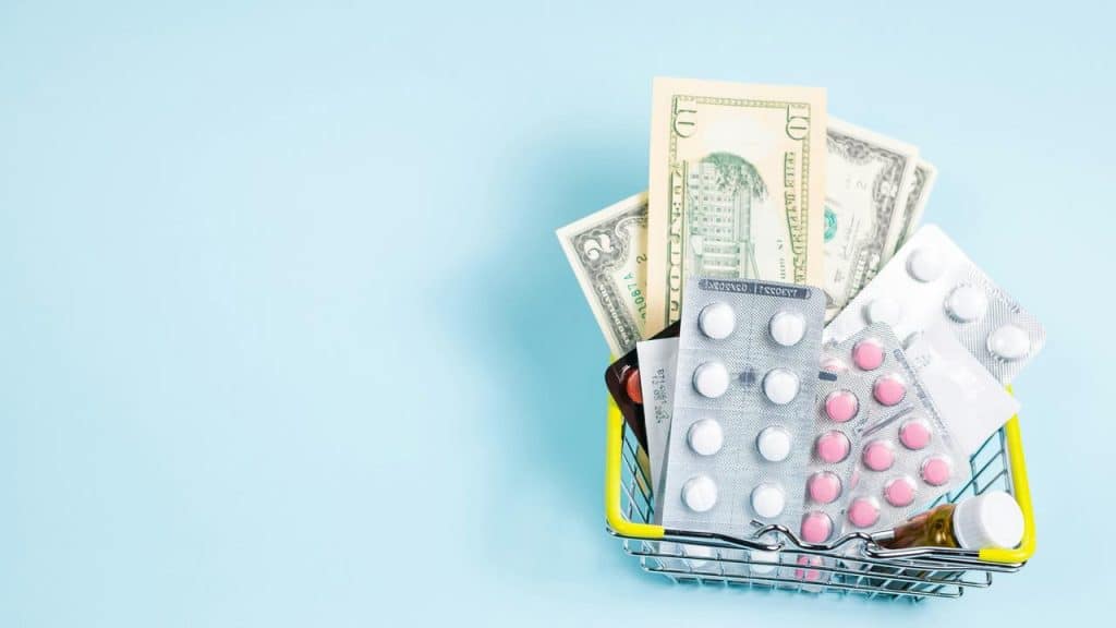 A small shopping basket holding blister packs of pills and U.S. dollar bills against a light blue background.