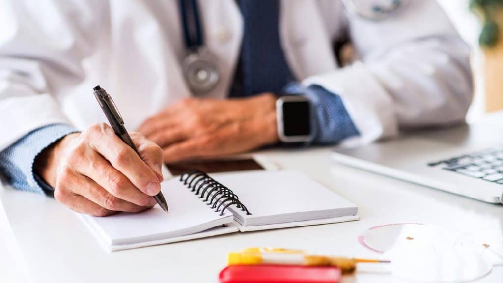 A doctor in a white coat writing in a spiral notebook at a desk.