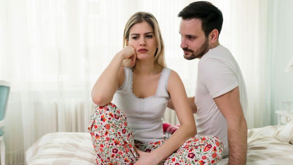 A man looks concerned at a woman sitting dejectedly in patterned pajamas.