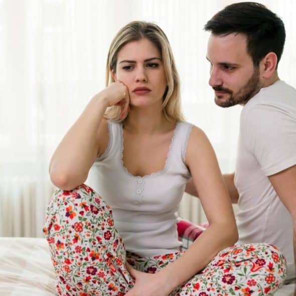 A man looks concerned at a woman sitting dejectedly in patterned pajamas.