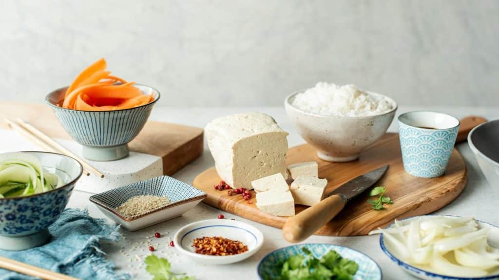 A block of cubed tofu on a wooden board with bowls of rice, vegetables, seeds, and spices.