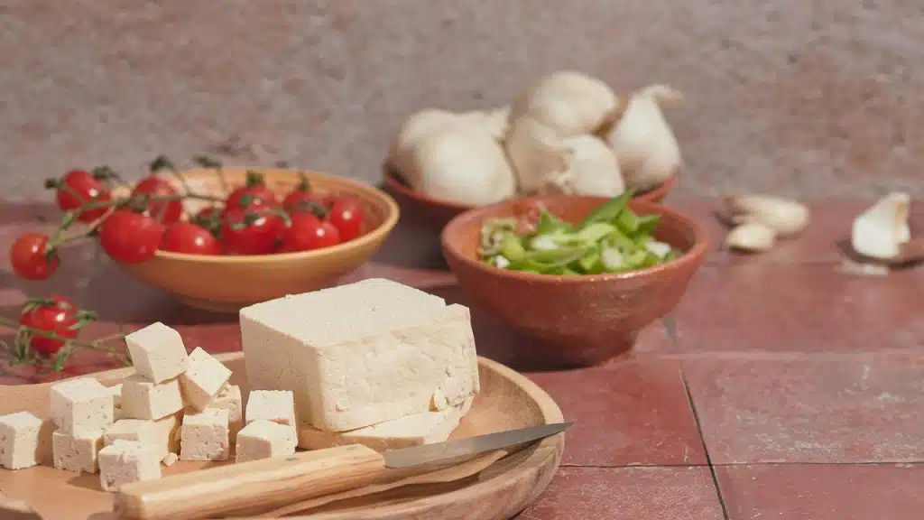 A block of cubed tofu on a board with bowls of cherry tomatoes and garlic.