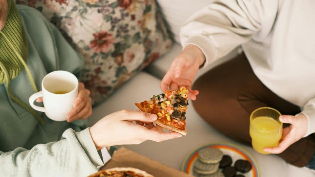 A hand passing a pizza slice between people holding drinks and cookies.