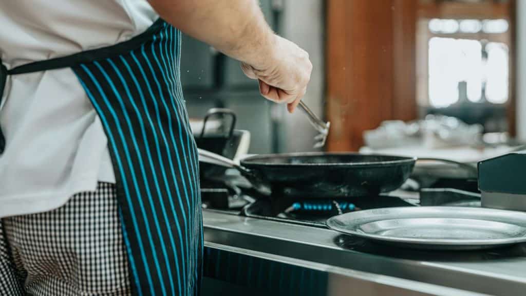 A cook in a striped apron stirring a skillet on a stovetop.