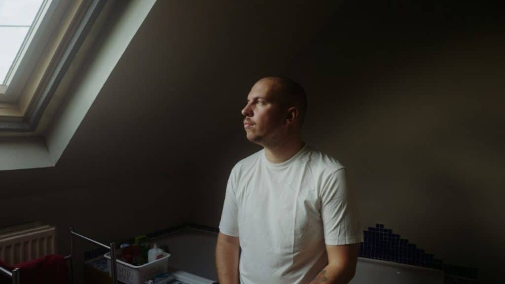 A bald man in a white t-shirt looks out a window in a dimly lit room.