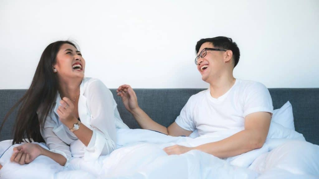 A happy Asian couple lies in bed, laughing and looking at each other.
