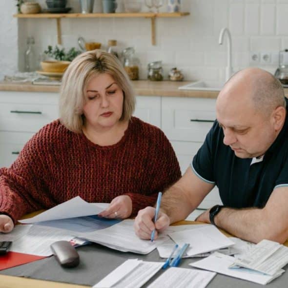 A couple sits at a kitchen table, reviewing documents and making calculations.