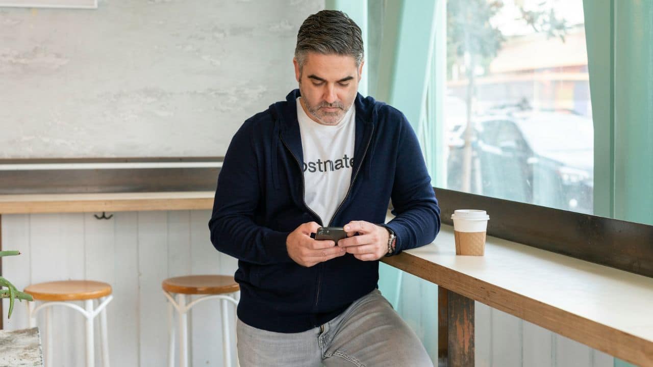 A man in a blue hoodie looks down at his phone, a coffee cup beside him.