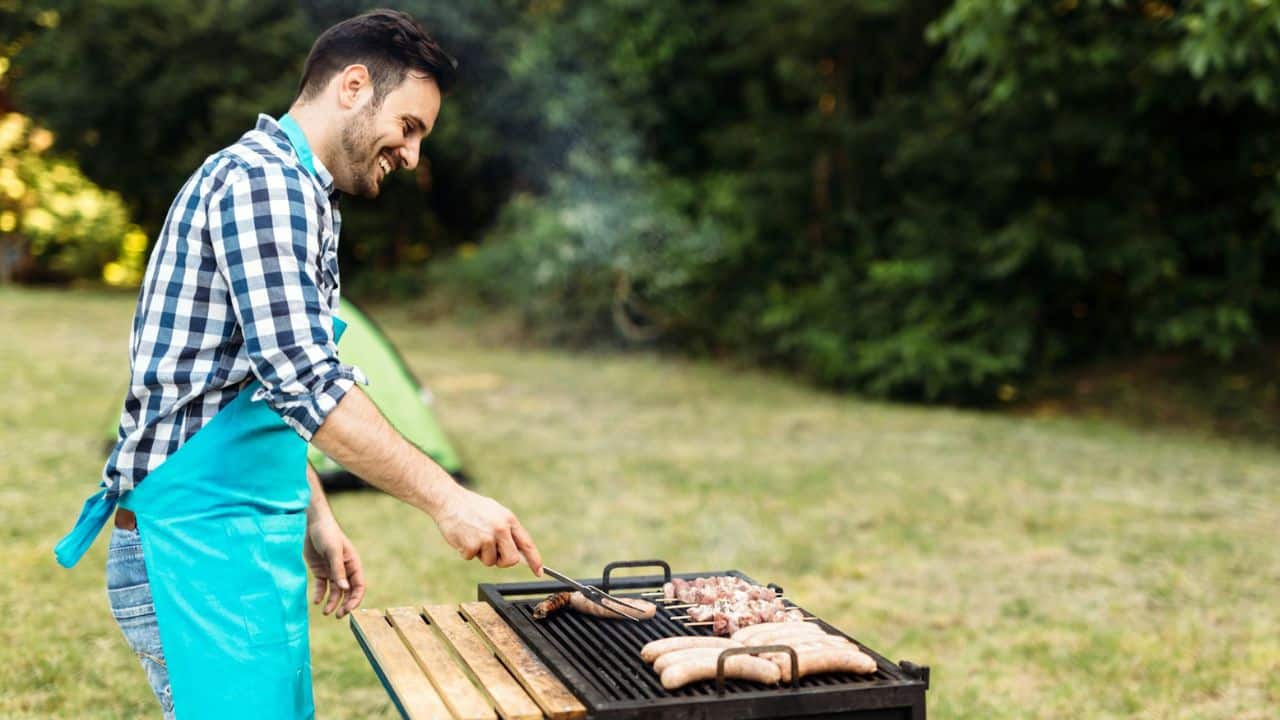 A smiling man in an apron grills sausages and meat outdoors.