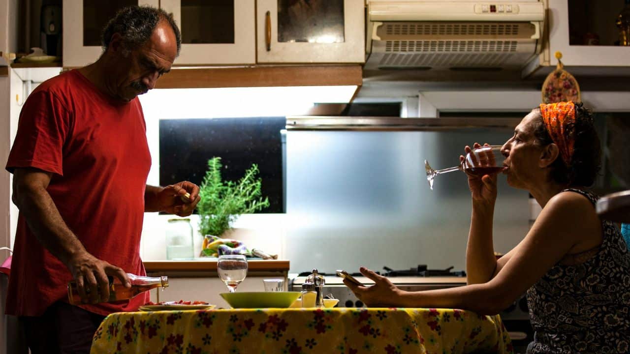 A man pours from a bottle while a woman sips wine at a kitchen table.