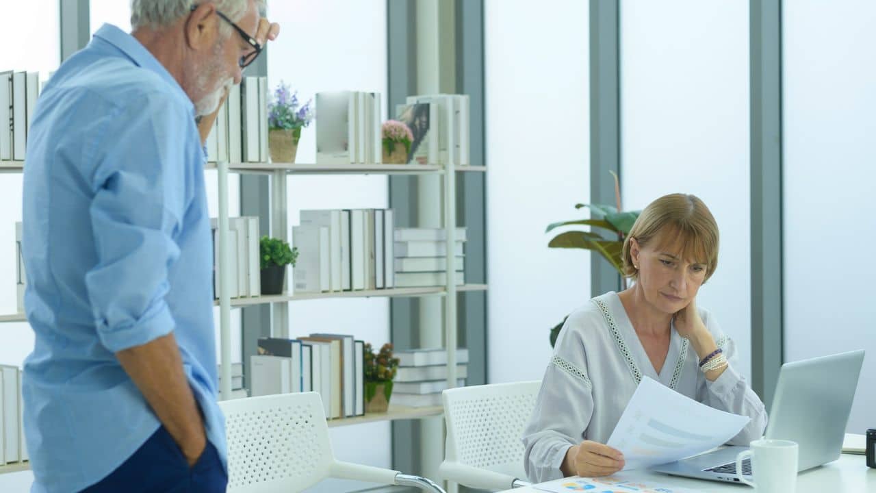 A concerned woman looks at papers while a man stands pensively in the background.