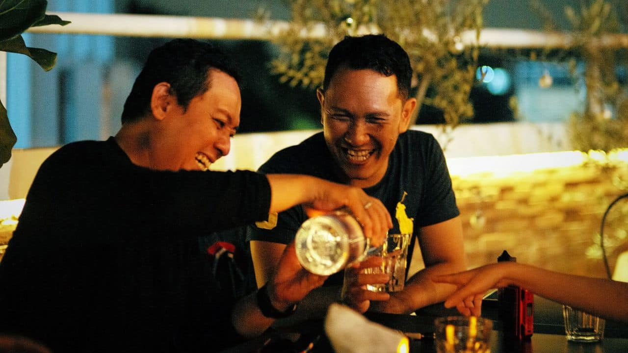 Two men are laughing heartily as one pours a drink for the other.