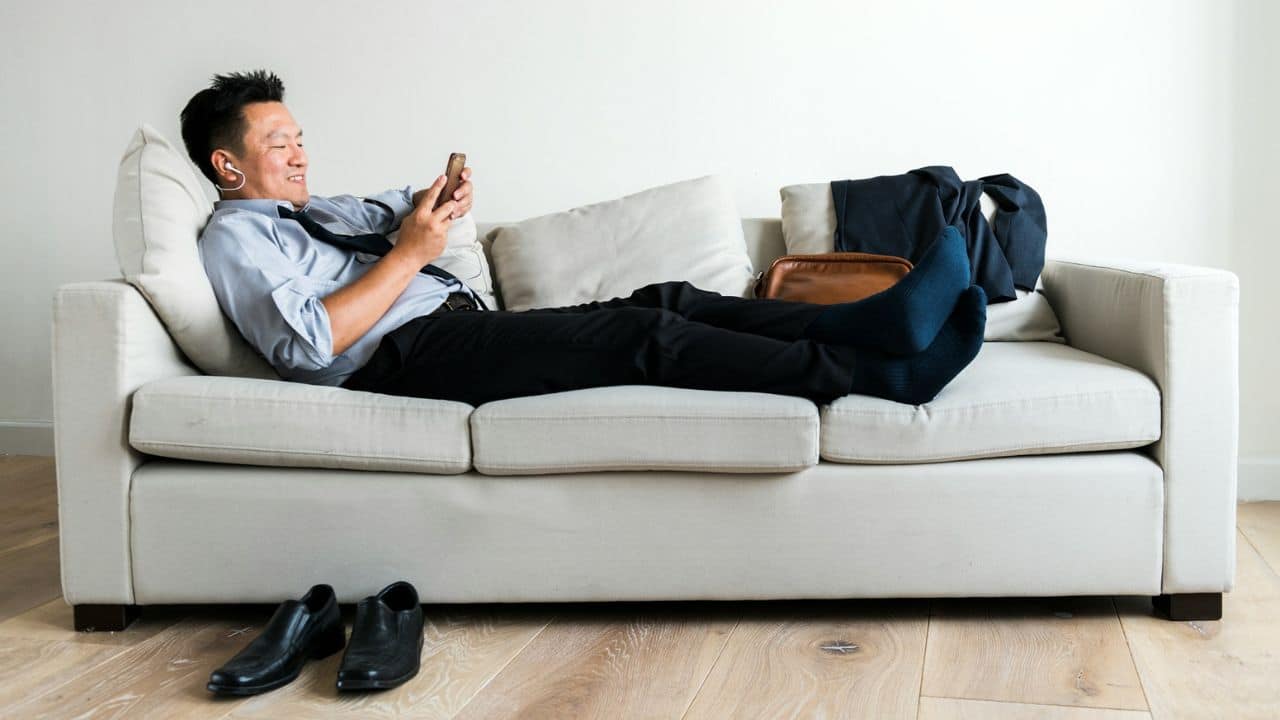 A man in a shirt and tie lies on a sofa, looking at his phone.