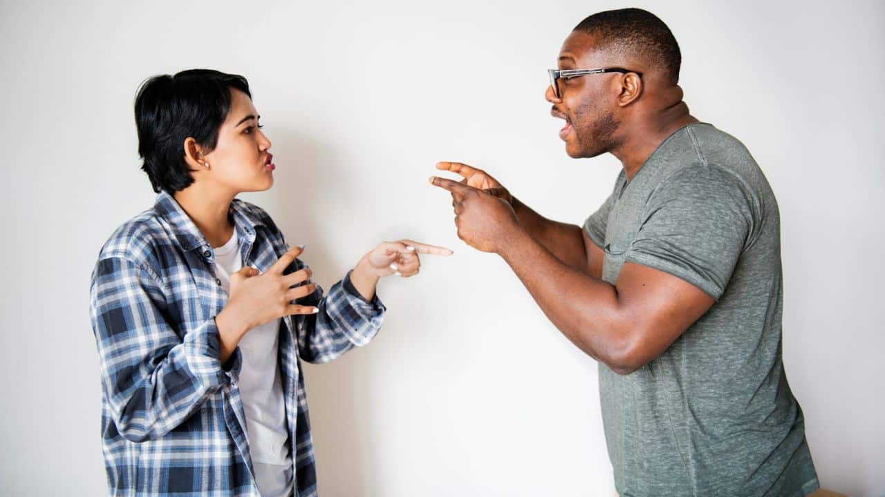 A couple argues, pointing fingers and expressing frustration in front of a white wall.