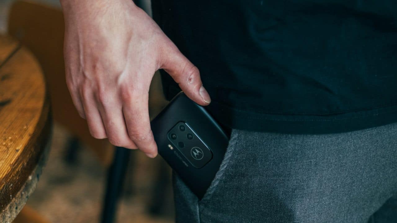 A person's hand slides a black Motorola phone with multiple cameras into a pants pocket.