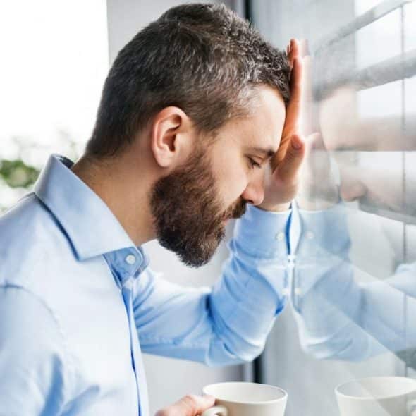 A man with a beard leans his head against a window, holding a mug.