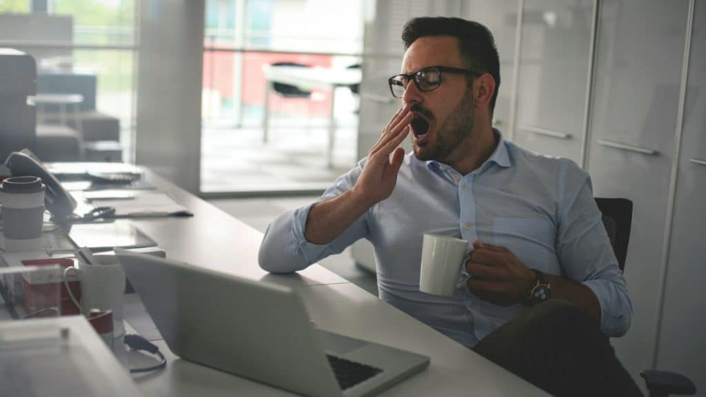 A man wearing glasses yawns and holds a mug while sitting at a desk with a laptop.