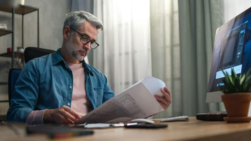 A grey-haired man with glasses reads a document at a desk with a computer monitor.