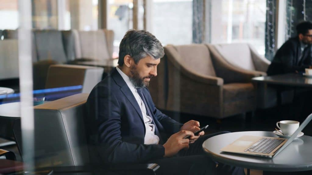 A bearded man in a suit looks at his phone while sitting in a cafe with a laptop.