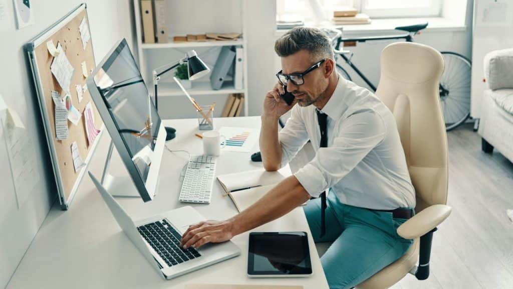 A man in a white shirt and glasses talks on a phone while working on a laptop at a desk.