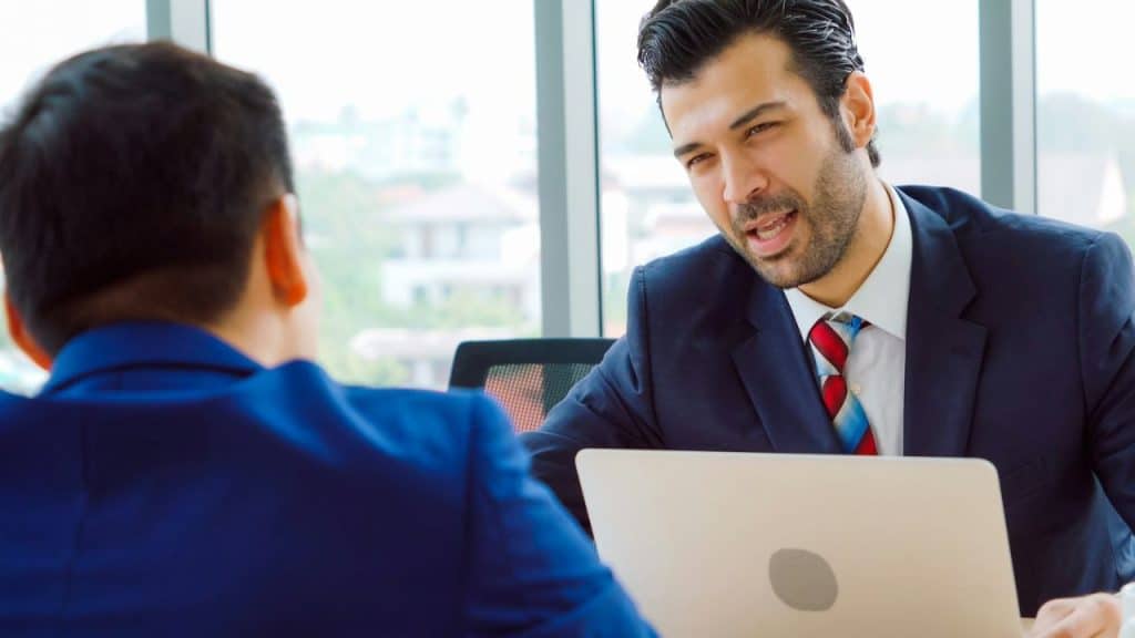 A man in a suit gestures with his hand while looking at another man.