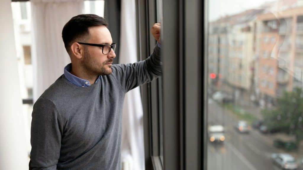 A man in glasses and a grey sweater looks out a window at a city street.
