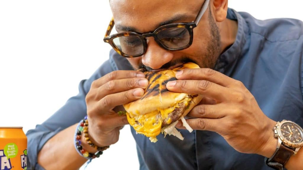A man with glasses and a beard takes a large bite out of a cheeseburger.