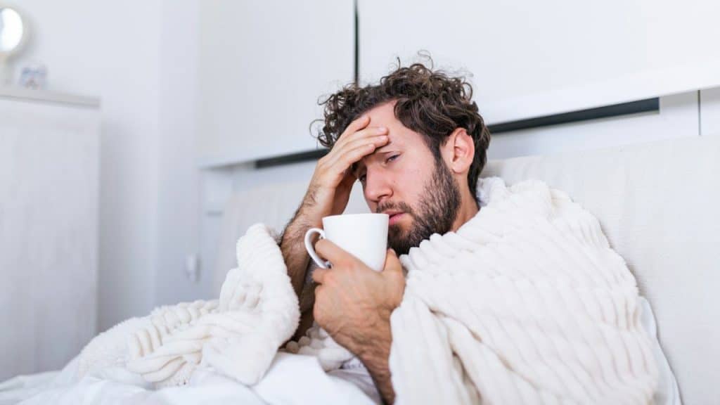 A man with curly hair, wrapped in a blanket, holds a mug and touches his forehead.