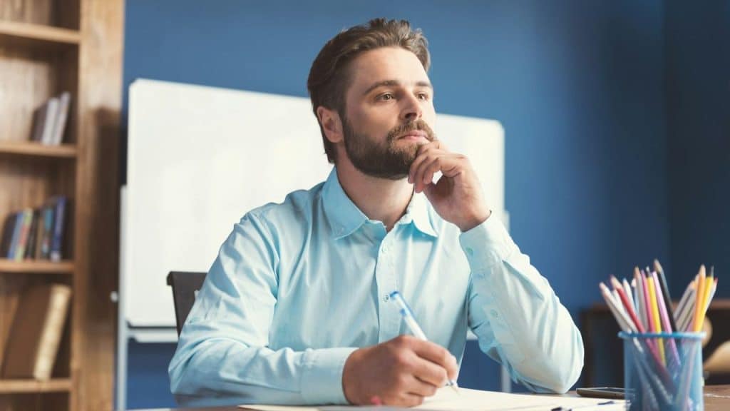 A bearded man in a light blue shirt sits at a desk, looking thoughtfully to the side.