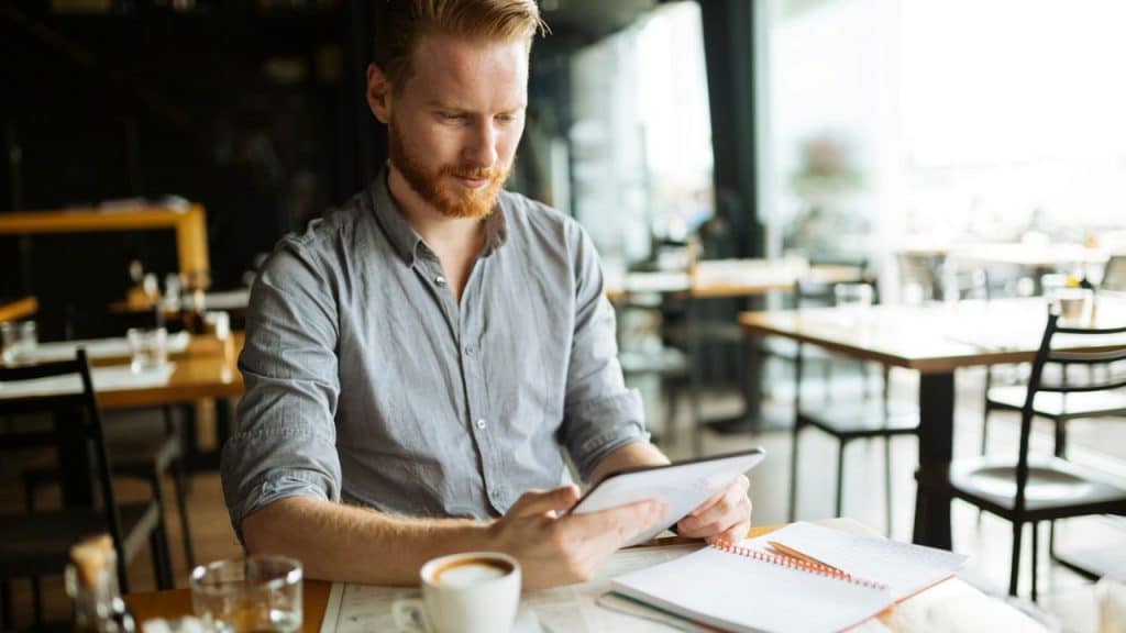 A red-bearded man in a grey shirt looks at a tablet at a table with coffee and a notebook.