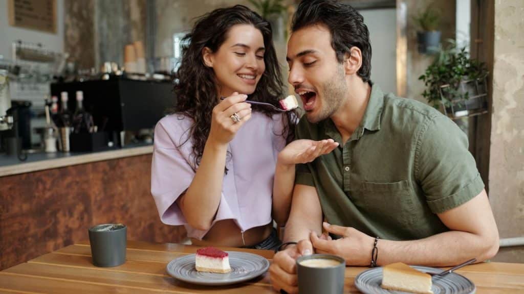 A woman feeds a piece of cake to a man, both smiling at a cafe table.