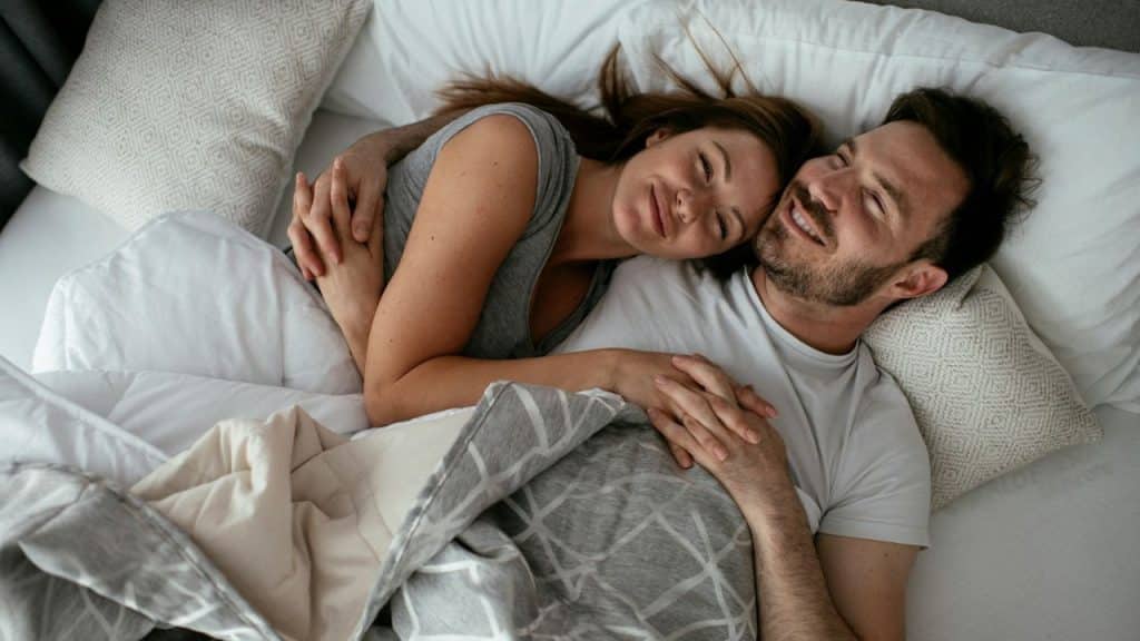 A happy couple is embraced in bed, both smiling and looking content.