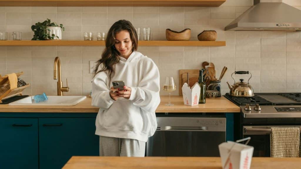 A young woman in a white hoodie smiles while looking at her phone in a modern kitchen.