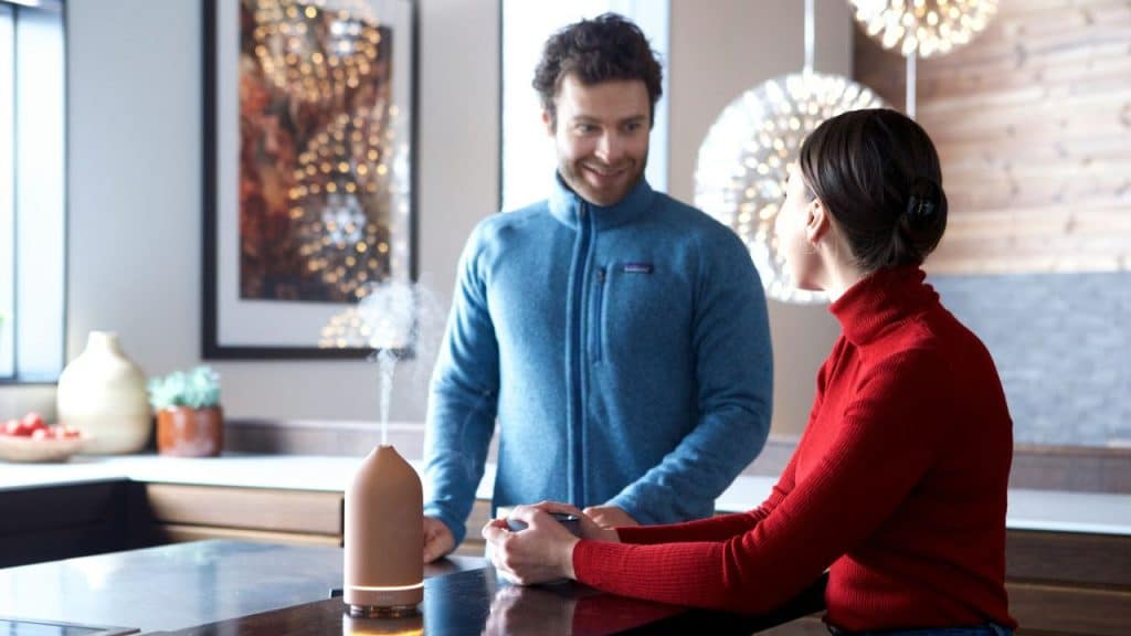 A man in a blue fleece smiles at a woman in a red turtleneck, with a diffuser between them.